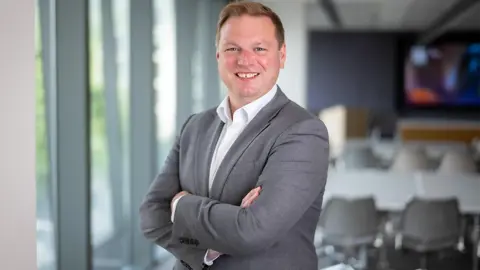 James Dand, Chief Operating Officer, in an office with a table, chairs and a large screen, and windows 