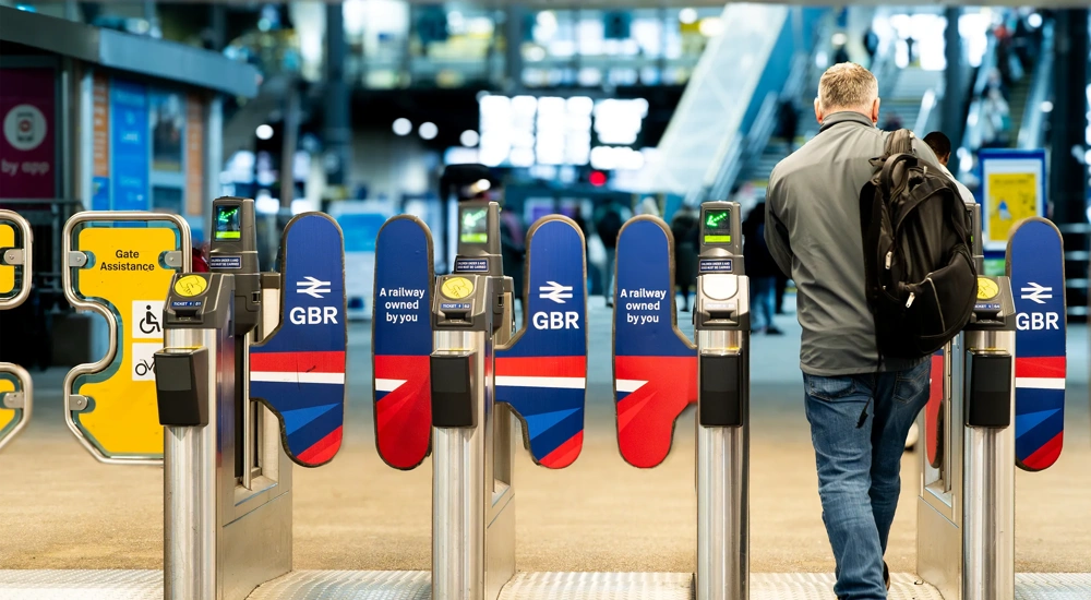 A rail passenger going through the ticket barrier at Leeds Train Station with the UK Government's re-nationalised rail company GBR signs and symbols displayed