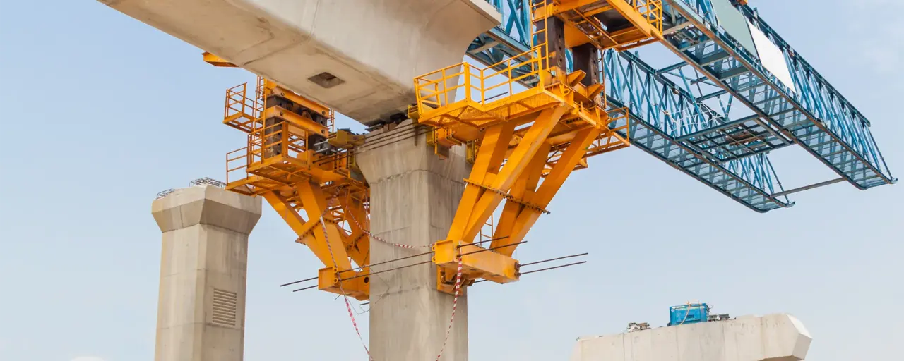 A large yellow and blue bridge construction crane is positioned above concrete support pillars, actively building a segmental bridge. The structure shows a partially completed bridge span with support equipment and materials on the site