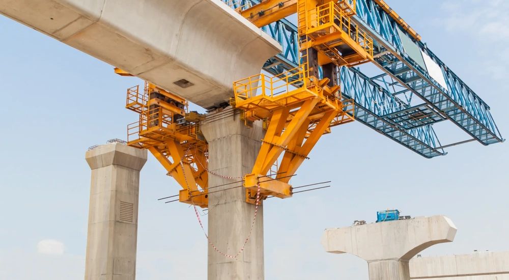 A large yellow and blue bridge construction crane is positioned above concrete support pillars, actively building a segmental bridge. The structure shows a partially completed bridge span with support equipment and materials on the site