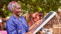 Woman wearing a purple Barefoot College International shirt holding a solar panel outside, in front of trees