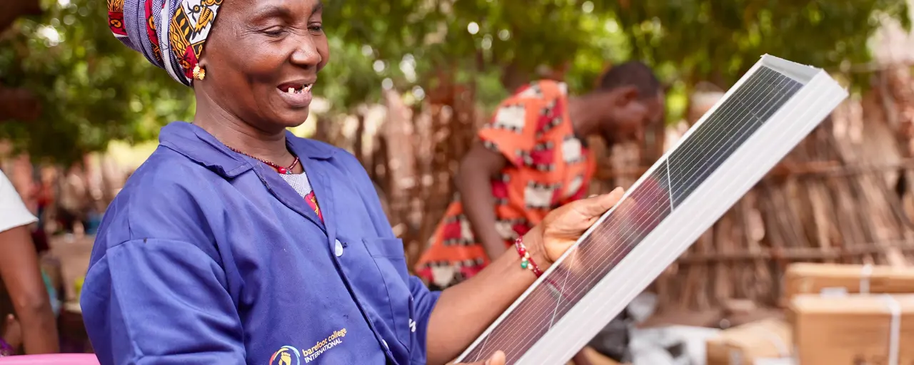 Woman wearing a purple Barefoot College International shirt holding a solar panel outside, in front of trees