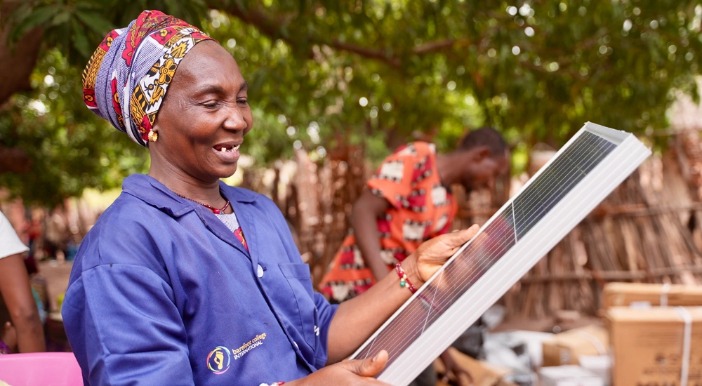 Woman wearing a purple Barefoot College International shirt holding a solar panel outside, in front of trees