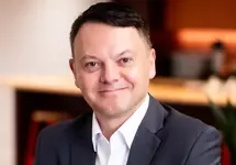 Portrait of Peter McGettrick, Managing Director, Advisory, standing in office kitchen space against light brown chair.