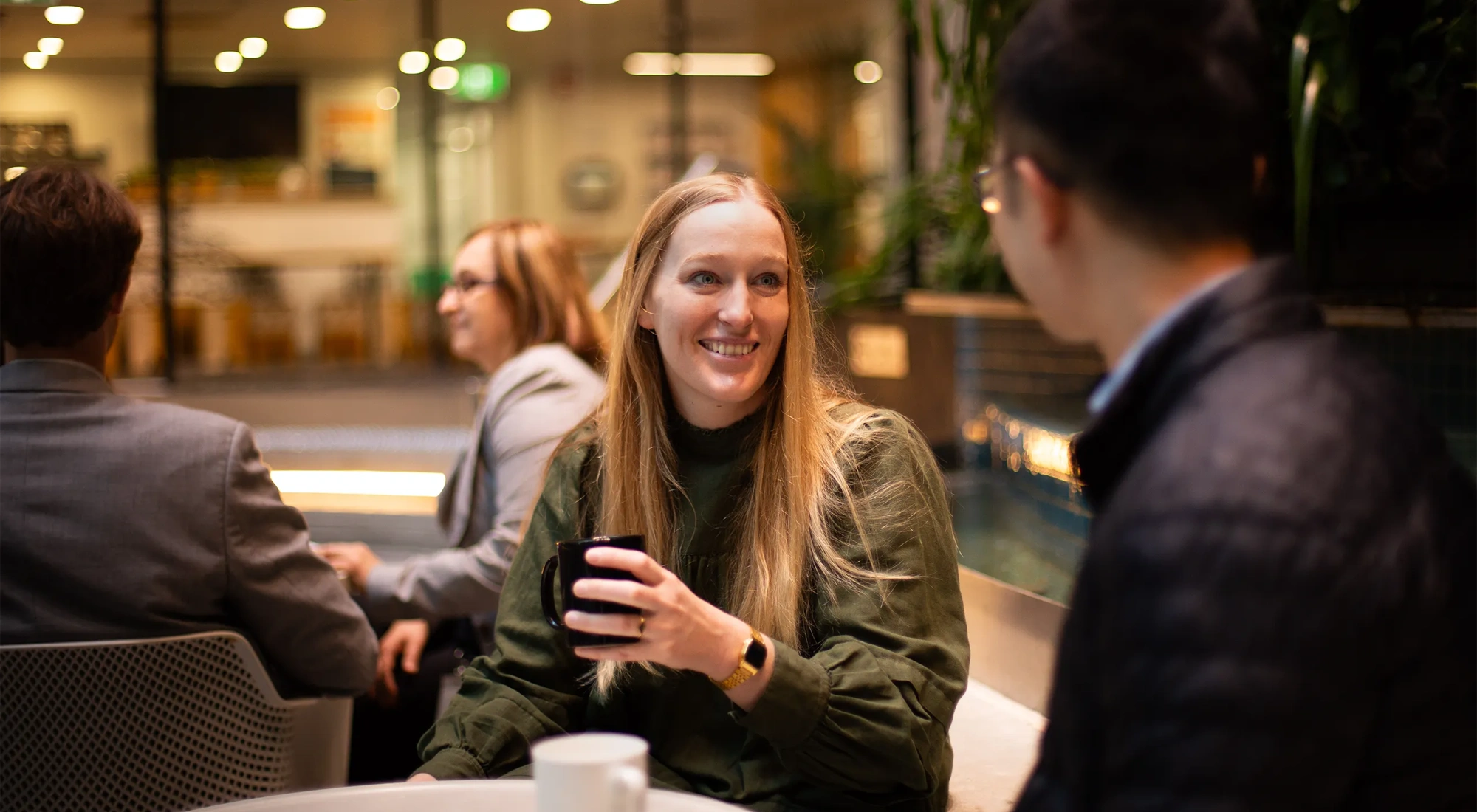 Two colleagues together shown from the males perspective, having a conversation in a plant-filled office, with blonde female colleague featured and smiling.