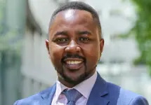 Robert Gichohi, Country Director, standing outside in front of an outdoor urban setting, featuring modern architectural elements with clean lines. There are hints of greenery, suggesting some trees or plants.