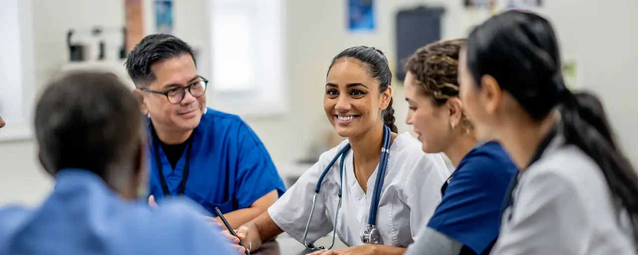 A small medical staff team sit around a boardroom table as they meet to discuss and collaborate on patient cases. They are each dressed professionally and are focused on the meeting.