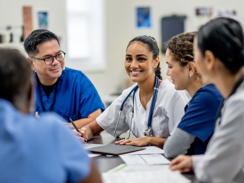 A small medical staff team sit around a boardroom table as they meet to discuss and collaborate on patient cases. They are each dressed professionally and are focused on the meeting.