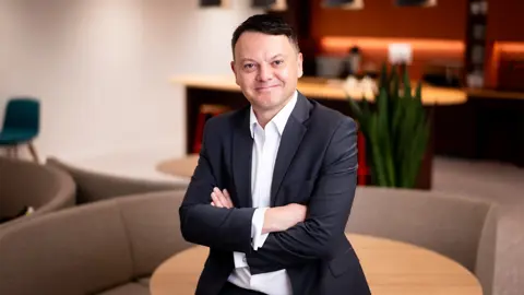 Portrait of Peter McGettrick, Managing Director, Advisory, standing in office kitchen space against light brown chair.