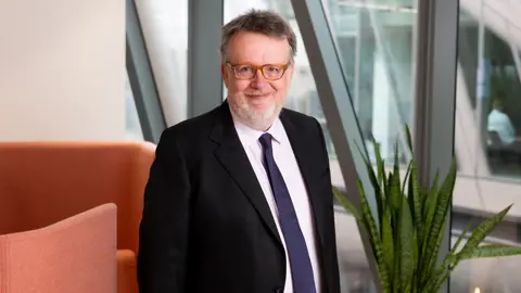 Portrait of Chris Hillier, Projects Expert, UK, standing in office space against orange chair.