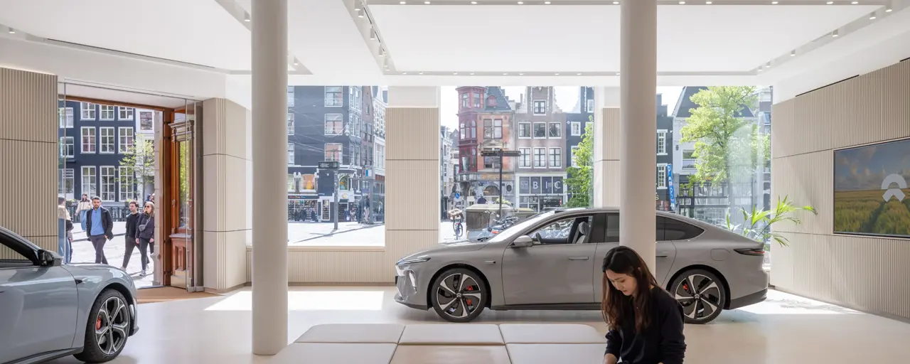 Interior of NIO gallery ground floor showroom. Two cars and a woman at a laptop feature in front of a glass backdrop of Amsterdam