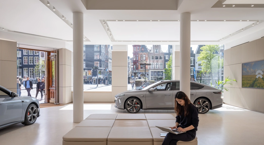 Interior of NIO gallery ground floor showroom. Two cars and a woman at a laptop feature in front of a glass backdrop of Amsterdam