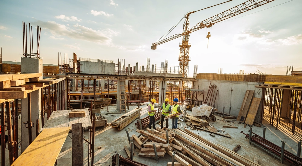 Three male construction workers discussing plans on the floor of a building under construction with a crane in background skyline.