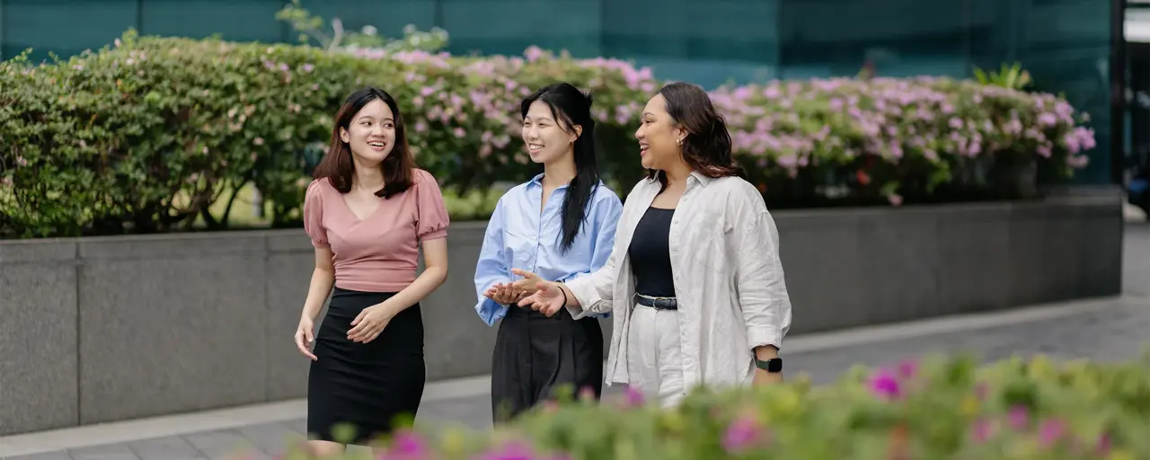 Group of three female colleagues walking and having a conversation outside surrounded by plants.