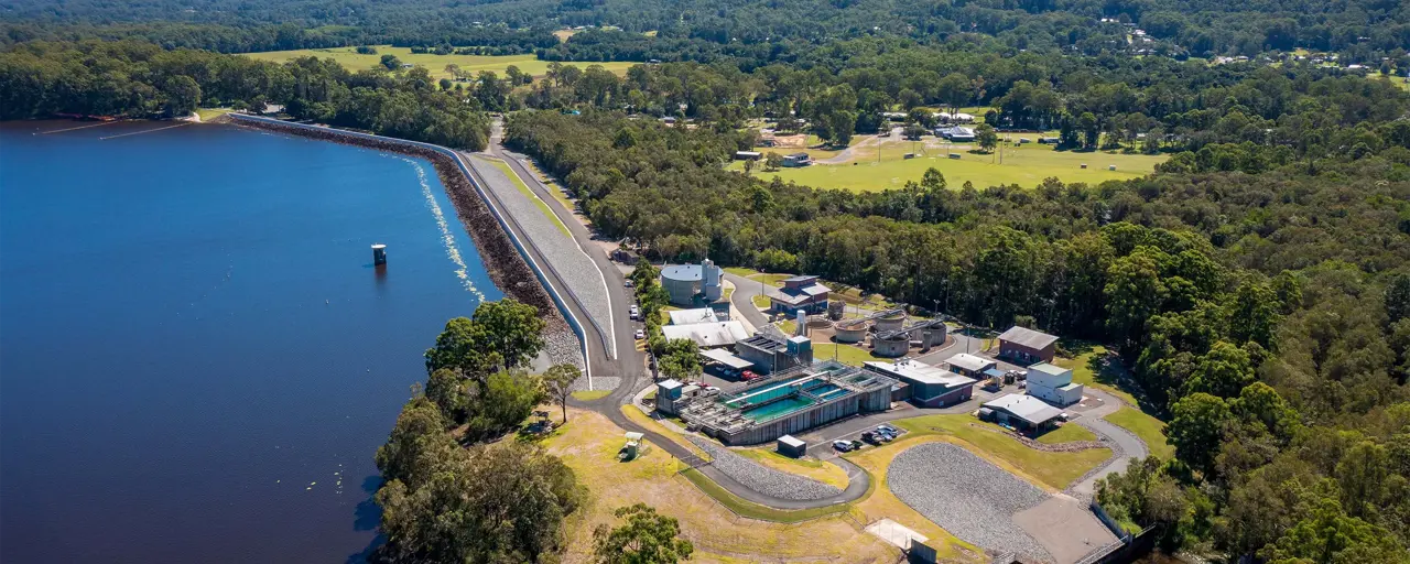 aerial view of a water treatment facility situated beside a large body of water, surrounded by dense green forested areas and rolling hills. The facility consists of several buildings, tanks, and infrastructure, positioned near a long, straight road running parallel to the water. The landscape includes a mix of forested and cleared areas with a few scattered buildings and open fields in the background.