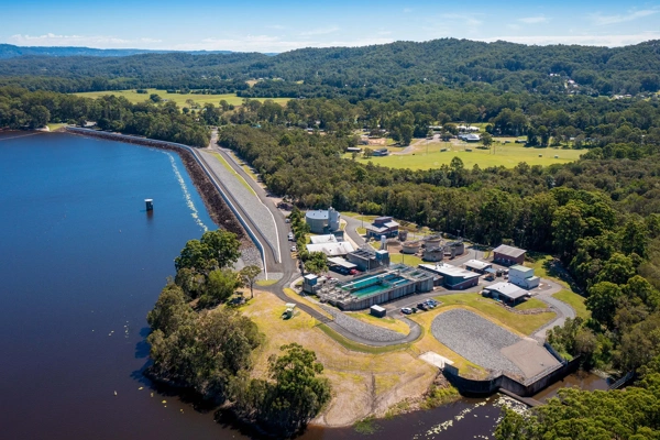 aerial view of a water treatment facility situated beside a large body of water, surrounded by dense green forested areas and rolling hills. The facility consists of several buildings, tanks, and infrastructure, positioned near a long, straight road running parallel to the water. The landscape includes a mix of forested and cleared areas with a few scattered buildings and open fields in the background.