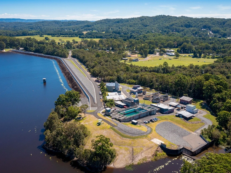aerial view of a water treatment facility situated beside a large body of water, surrounded by dense green forested areas and rolling hills. The facility consists of several buildings, tanks, and infrastructure, positioned near a long, straight road running parallel to the water. The landscape includes a mix of forested and cleared areas with a few scattered buildings and open fields in the background.