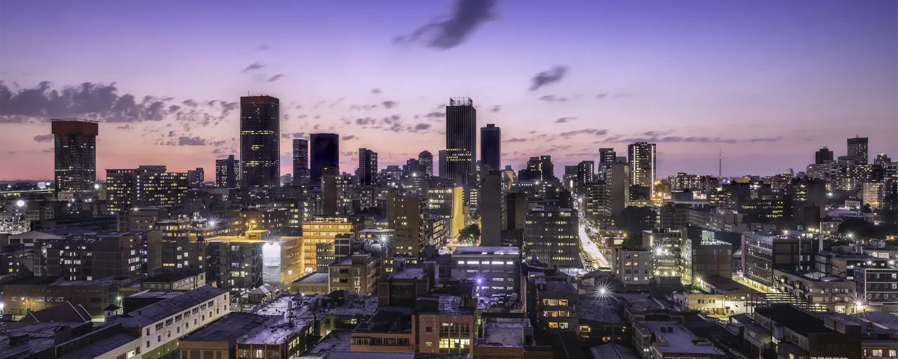 Image of city with tall buildings filled with lights, against blue and purple sunset