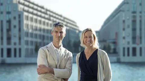 Portrait of Bettina Würfel, Managing director of Austria and Germany, and Stefan Reimoser, standing in front of modern urban architecture with two large, multi-story buildings featuring rows of windows on either side of a canal or river. The buildings are symmetrical and have a contemporary design, with a clear sky above and water reflecting the structures below.