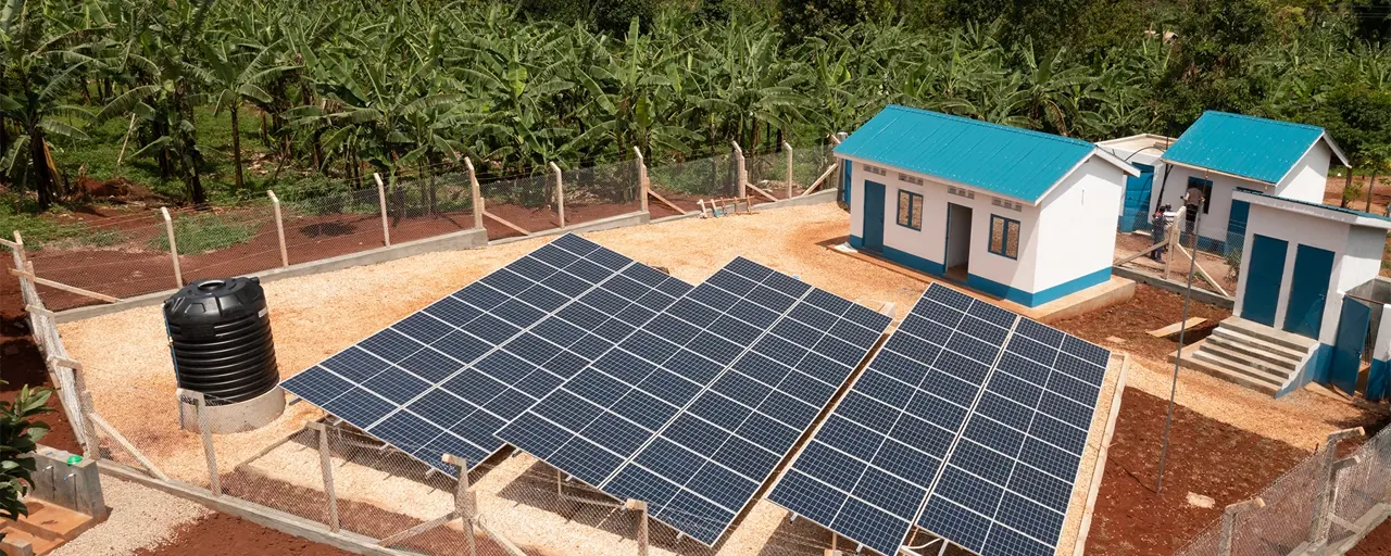 Three solar panels next to a few small houses with blue roofs, with a background of jungle foliage.