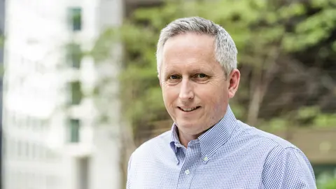 Martin Sudweeks, UK Managing Director, Cost and Commercial Management, smiling in a blue shirt outside in front of trees