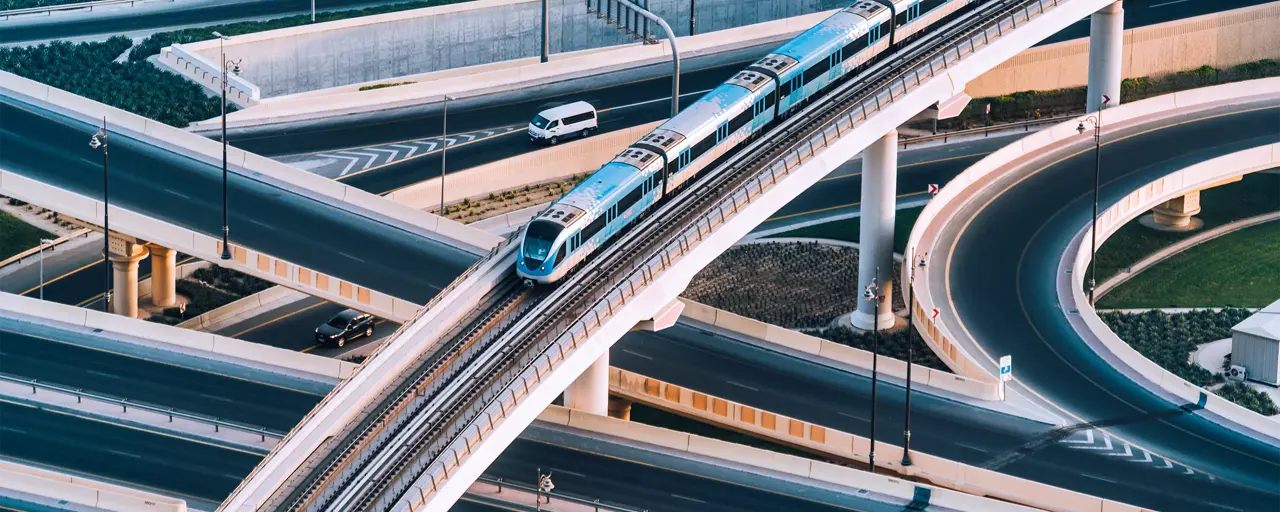 An elevated blue and white metro train travels along a curved, multi-level bridge over a network of highways. Several cars are visible on the roads below, navigating the complex system of interchanges. The image captures modern urban infrastructure, with smooth, sweeping curves and elevated tracks against a backdrop of landscaped areas and concrete structures