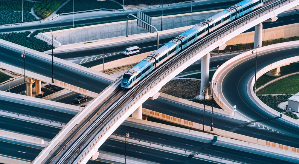 An elevated blue and white metro train travels along a curved, multi-level bridge over a network of highways. Several cars are visible on the roads below, navigating the complex system of interchanges. The image captures modern urban infrastructure, with smooth, sweeping curves and elevated tracks against a backdrop of landscaped areas and concrete structures
