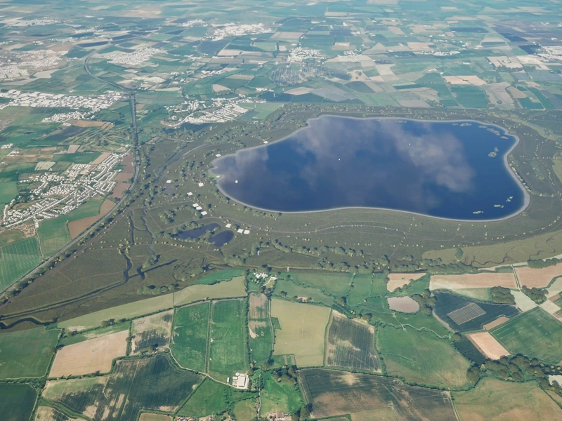 An aerial view of a large stone dam with a reservoir on the right side and a lush green landscape surrounding it. A small building is visible near the base of the dam, adjacent to a body of water and rocky terrain.