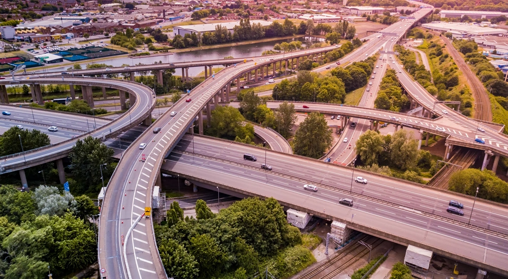 Aerial view of a complex motorway road junction with traffic moving. Cars, lorries, vans and a train can be seen travelling through a busy road interchange from above.