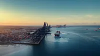 An aerial view of a large shipping port at sunset. The port is lined with numerous container cranes and stacks of shipping containers. A cargo ship is seen navigating out of the port towards the open sea, leaving a wake behind. The sky is clear with a gradient from light yellow to blue as the sun sets in the distance.