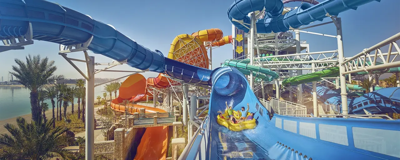 Big water park with lots of slides, featuring family on water float going down big blue water slide with heir arms raised in the air