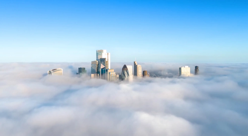 City skyline with modern skyscrappers rising above a layer of fog under a clear blue sky.