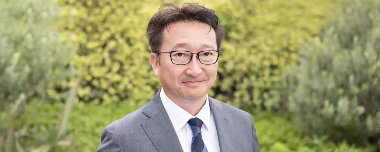 Portrait of Toshio Kosuge, director of clean energy and transmissions in Japan, standing outside against a green background