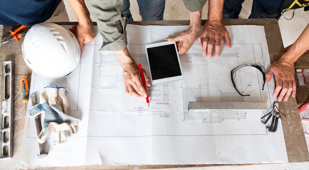 Three work people reading building plans on a table, includes iPad and construction work hat and safety glasses