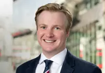 Portrait of George Highfield, Real Estate Advisory, Data & Digital – Principal Consultant, standing outside in front of buildings featuring an urban cityscape with a mix of architectural styles. The scene includes some windows and red features, adding a touch of color.