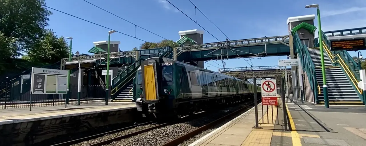 train station platform with a modern train passing through. Overhead electrified lines are visible, and there is a footbridge connecting both sides of the station. The station has green and white signage, and the platform area includes a yellow tactile paving strip for safety. Stairs with green railings lead up to the footbridge on either side, and a "No Entry" sign is visible near the platform edge. The sky is clear and blue, indicating a sunny day.