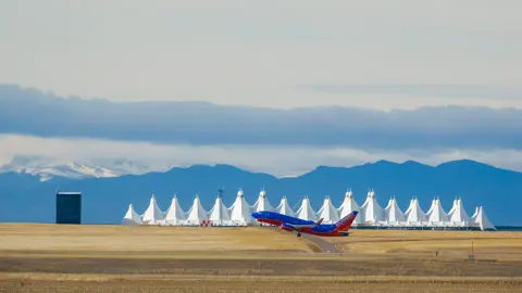  A blue and red aeroplane just taking off from the runway against a backdrop of the airport and snowcapped mountains. 