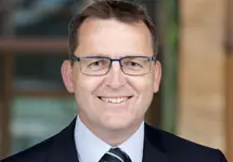 Portrait of Peter Coombs,  Director, outside in front of a blurred brown background featuring a building and mix of natural and urban elements.