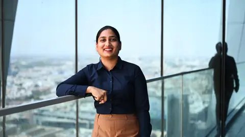 Priyanka Narne, Senior Cost Manager, smiling in front of a window with a large cityscape view of Riyadh