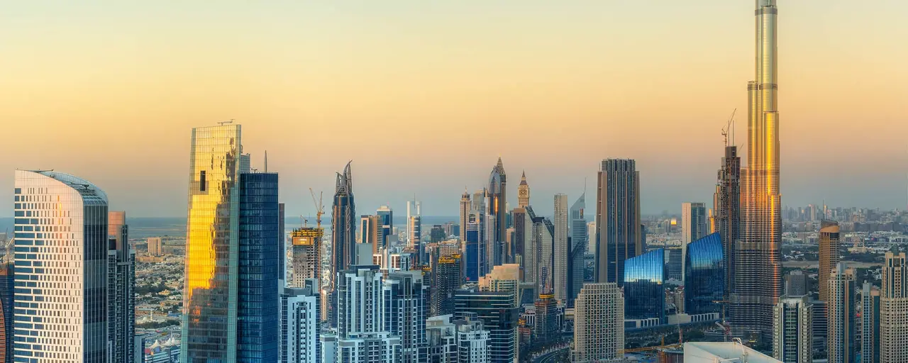 A panoramic view of a modern city skyline at sunset, featuring numerous high-rise buildings and skyscrapers, including the iconic Burj Khalifa on the right, dominating the landscape with its towering height. The buildings are illuminated by the warm golden light of the setting sun, creating a striking contrast against the clear sky.