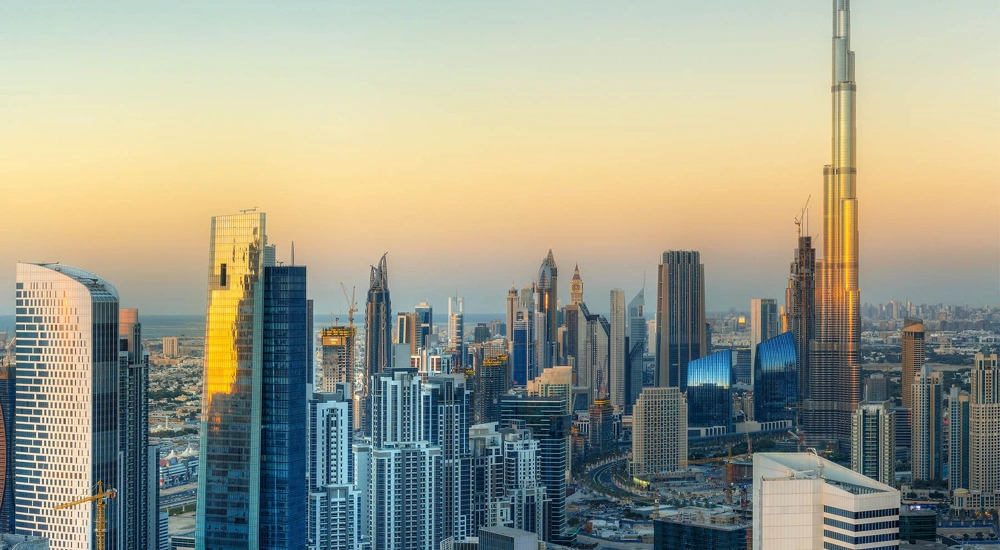A panoramic view of a modern city skyline at sunset, featuring numerous high-rise buildings and skyscrapers, including the iconic Burj Khalifa on the right, dominating the landscape with its towering height. The buildings are illuminated by the warm golden light of the setting sun, creating a striking contrast against the clear sky.