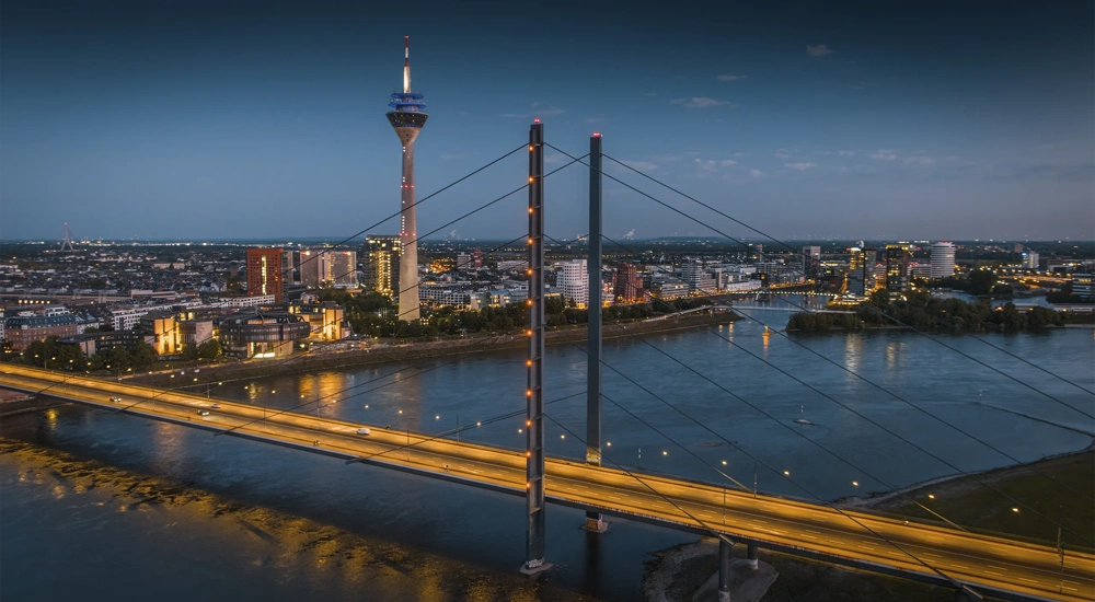 Aerial blue-hour view of the Düsseldorf skyline along the Rhine River, featuring the illuminated Rheinkniebrücke leading toward the city center and the Rheinturm rising above modern buildings. City lights reflect on the calm water, creating a clean, contemporary European cityscape. North Rhine–Westphalia, Germany, Europe.