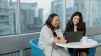 Two female colleagues gathered around table sitting on blue chairs looking at laptop.