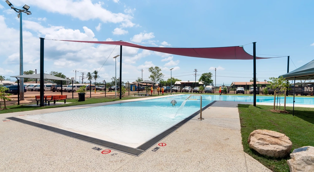 An outdoor swimming pool with shallow water, surrounded by a concrete deck. The pool has a sloped entry and a handrail on the right side. There are signs on the pool deck indicating "No Diving" and depth markings in both feet and meters. Above the pool, a large red sunshade is stretched between several poles to provide shade. The surrounding area includes some green grass, benches, and small trees. In the background, there are buildings, palm trees, and parked cars under a partly cloudy sky.