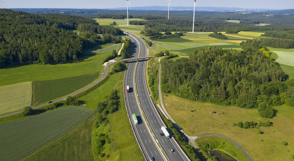 Traffic on a motorway surrounded by green fields, forest and wind turbines in the far distance