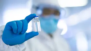 A person dressed in protective laboratory clothing, including blue latex gloves and holding vaccination vile in between their fingers. 