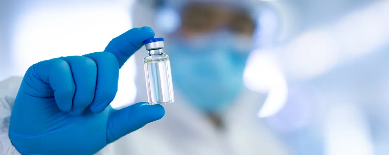 A person dressed in protective laboratory clothing, including blue latex gloves and holding vaccination vile in between their fingers. 