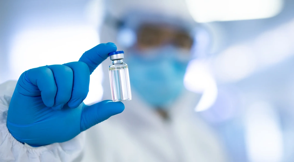 Lab technician in protective white suit, face mask, goggles and blue gloves holding a glass vial