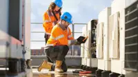 Two workers in orange safety jackets and blue helmets performing maintenance on HVAC units. One worker is kneeling and using a multimeter to check electrical connections, while the other worker is standing behind, observing and taking notes. The setting appears to be a rooftop with buildings visible in the background.