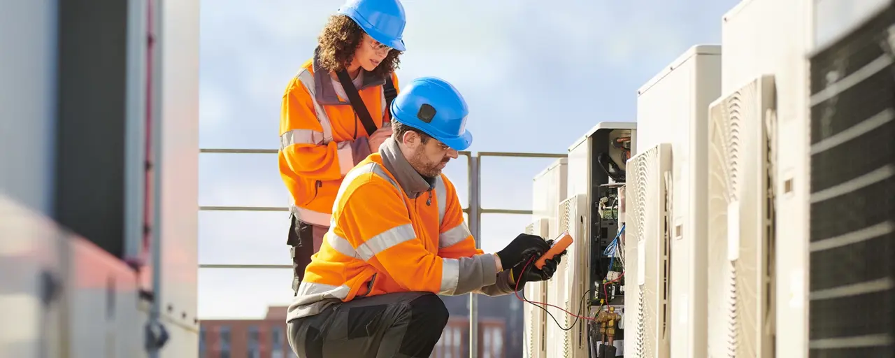 Two workers in orange safety jackets and blue helmets performing maintenance on HVAC units. One worker is kneeling and using a multimeter to check electrical connections, while the other worker is standing behind, observing and taking notes. The setting appears to be a rooftop with buildings visible in the background.